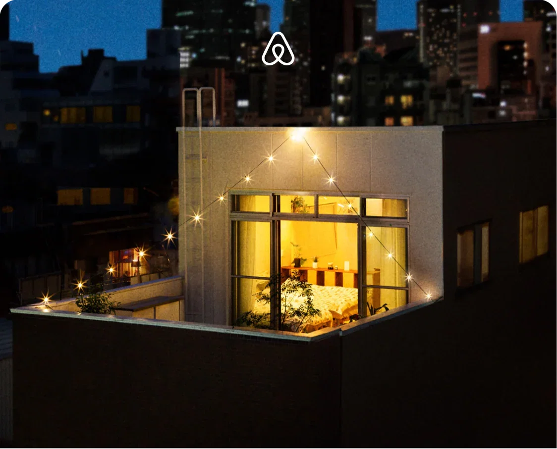 Cozy rooftop apartment glowing with warm light at night, surrounded by city buildings and string lights.