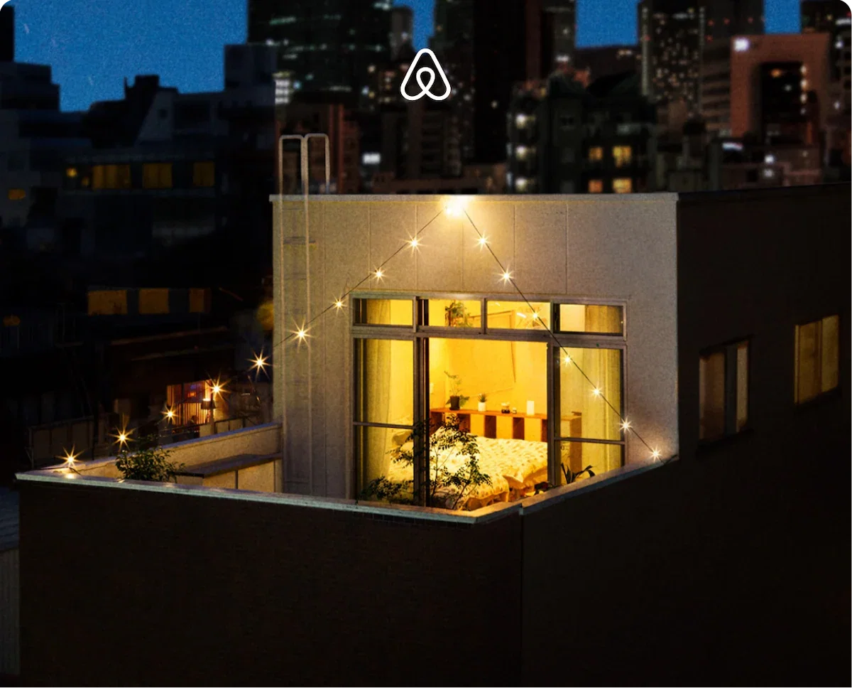 Cozy rooftop apartment glowing with warm light at night, surrounded by city buildings and string lights.