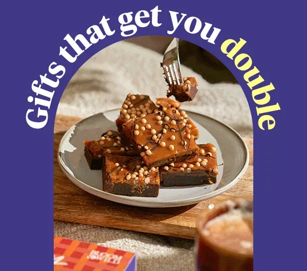 Stack of rich chocolate brownies topped with glossy caramel, chocolate drizzle and crunchy biscuit pearls on a ceramic plate. A fork lifts a fudgy bite. Arched text reads 
