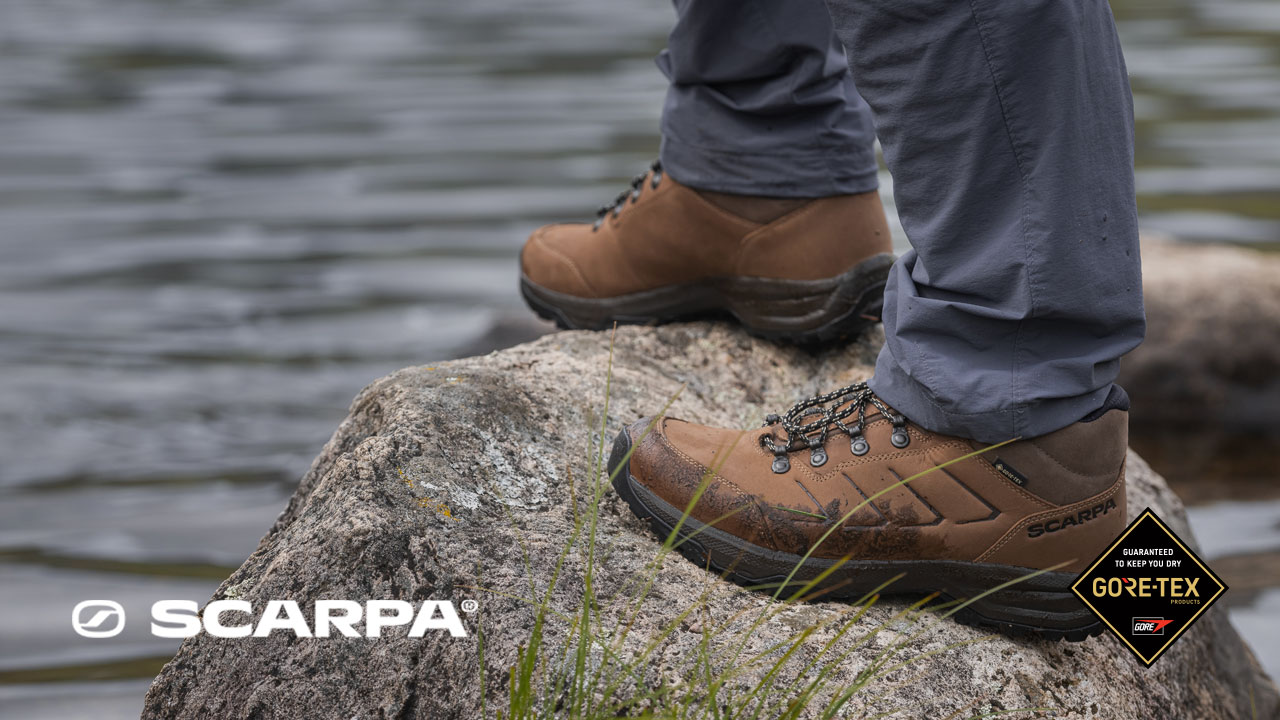 Close-up of brown leather hiking boots standing on a rock beside a rippling lake, with grey outdoor trousers visible and soft water textures in the background.