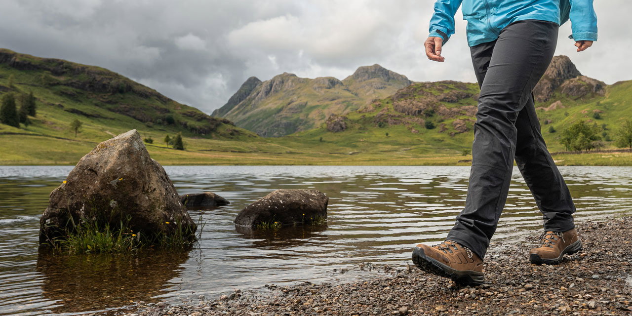 Outdoor scene of a person wearing a blue waterproof jacket, grey trousers, and brown hiking boots walking along a lakeshore, with large rocks in the water and green hills under a cloudy sky in the background.