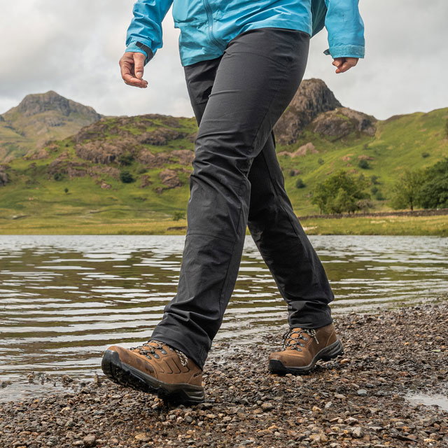 Outdoor scene of a person wearing a blue waterproof jacket, grey trousers, and brown hiking boots walking along a lakeshore, with large rocks in the water and green hills under a cloudy sky in the background.
