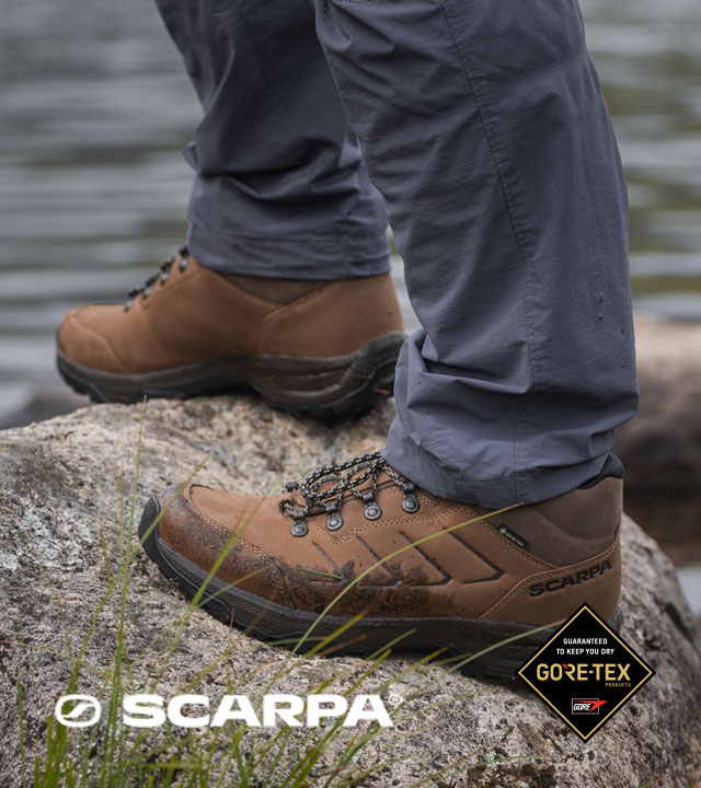 Close-up of brown leather hiking boots standing on a rock beside a rippling lake, with grey outdoor trousers visible and soft water textures in the background.
