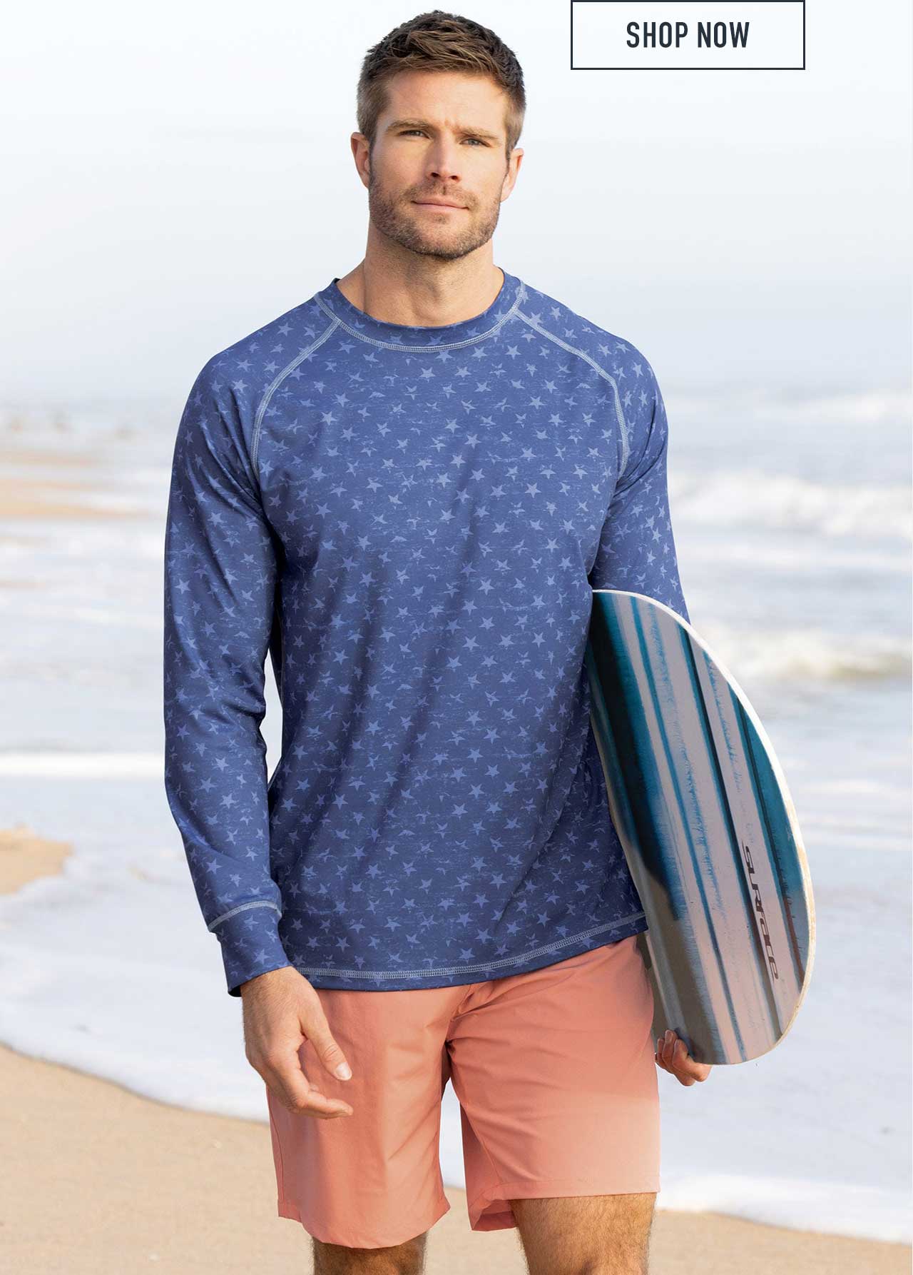 Man standing on beach holding a surfboard, wearing a blue long-sleeve rash guard with all-over star print and coral swim shorts, with Shop Now button