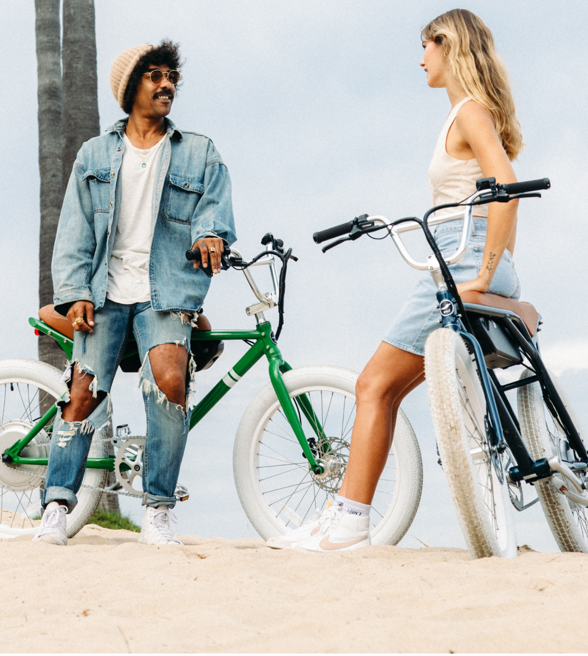 Two people chatting on a beach with bicycles, wearing casual outfits.