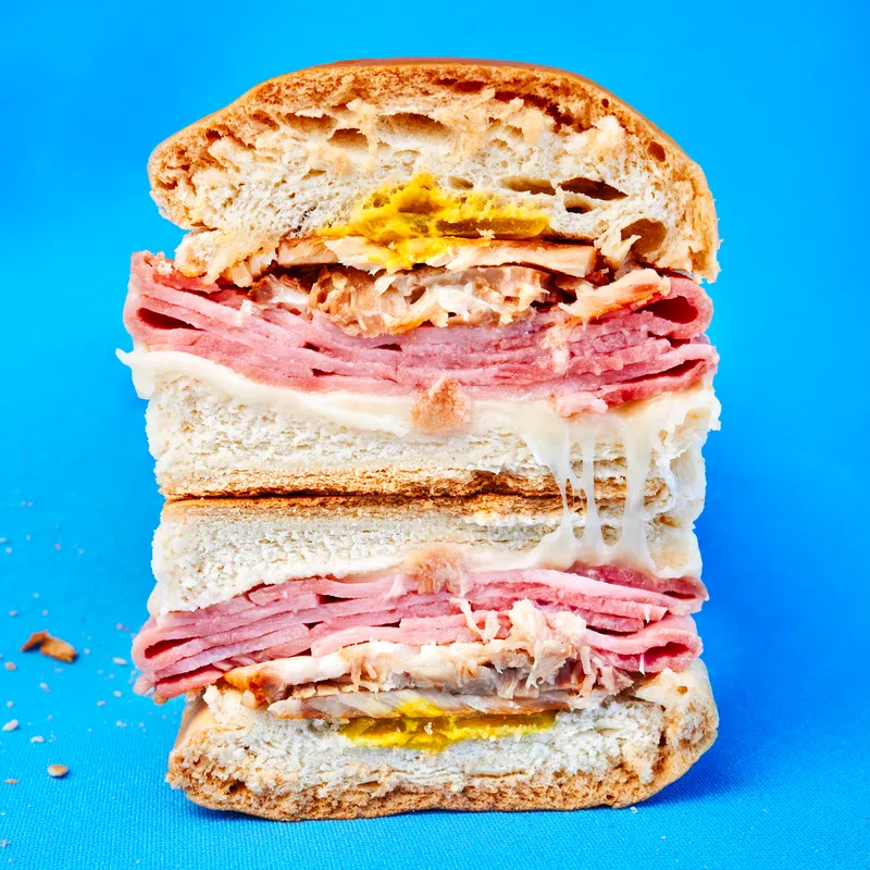 Two halves of a Cuban sandwich, connected by a stringy cheese pull, on a vibrant blue background. 