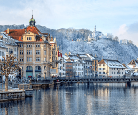 Snow-covered buildings along a calm lake, with a castle on a hill in the background.