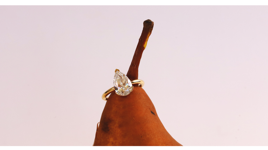 Close up of pear diamond engagement ring on pear stem, against a light pink background.