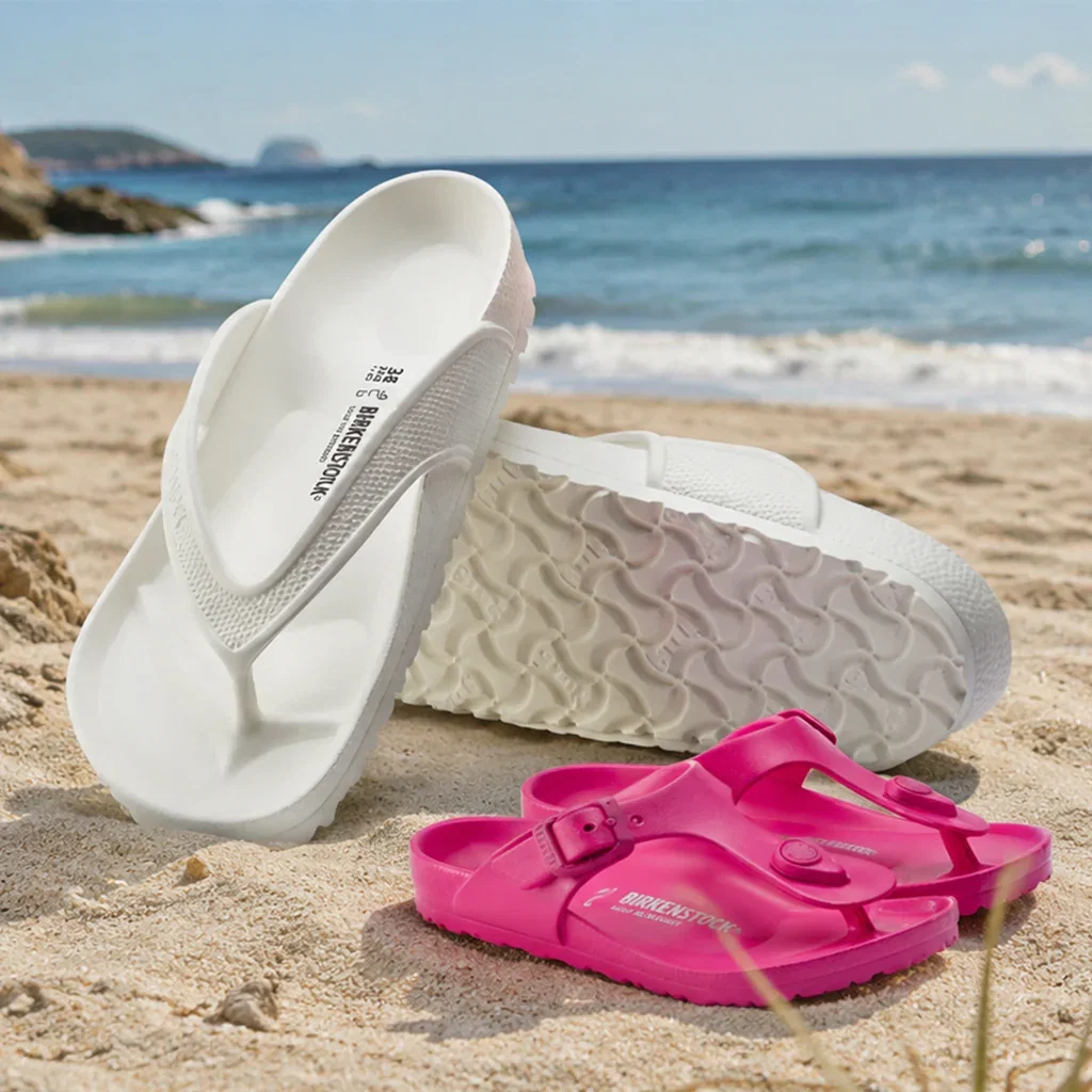 Birkenstock EVA sandals in white and pink displayed on sandy beach with ocean waves in background.