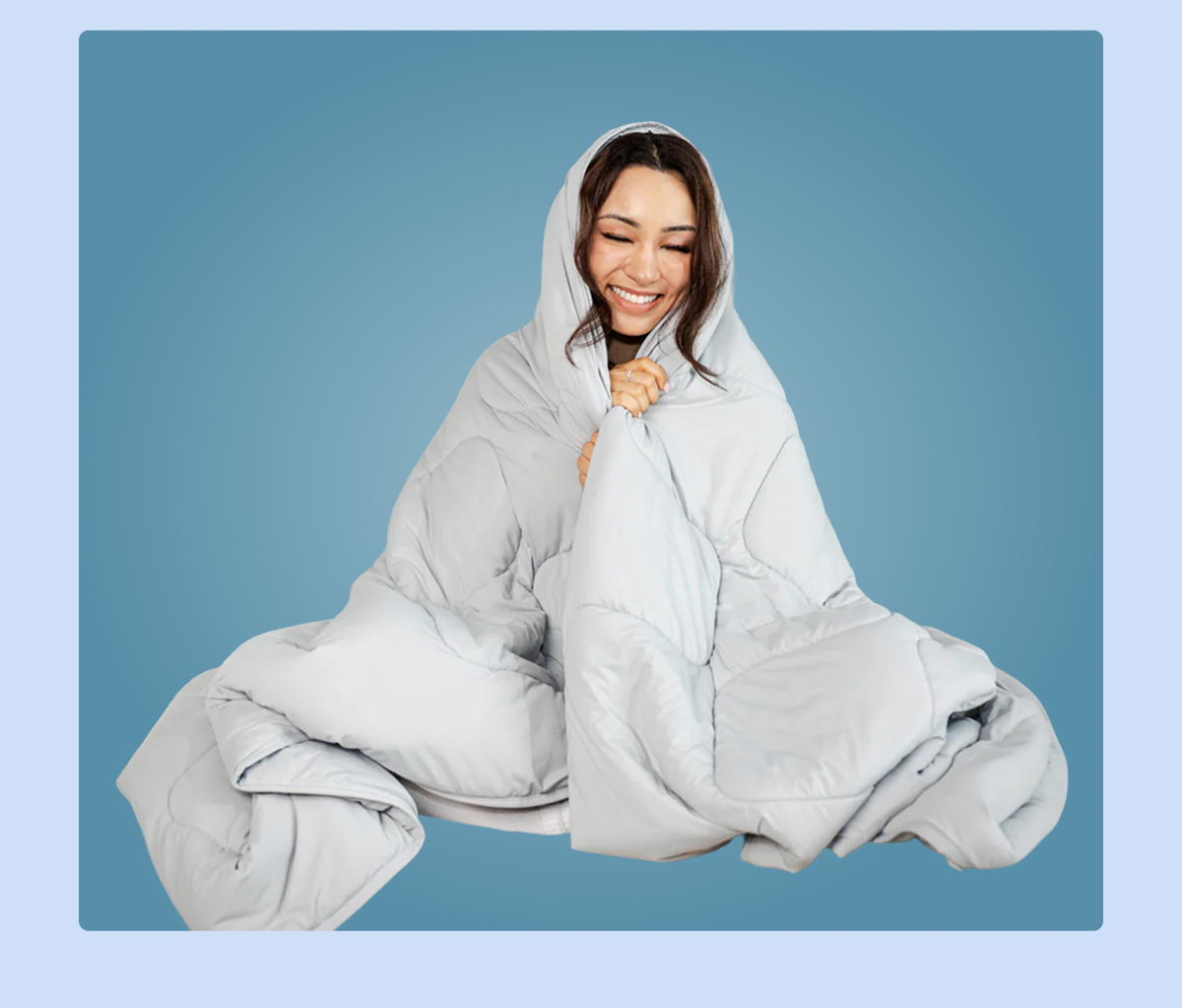 A woman smiling while wrapped in a cozy gray blanket against a blue background.