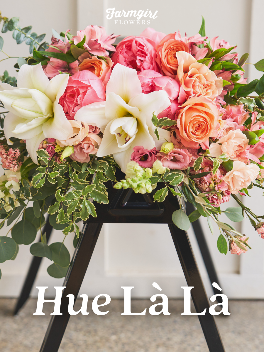 A lush Farmgirl Flowers bouquet on a black easel, full of pink peonies, coral roses, white lilies, and trailing greenery, with the words 