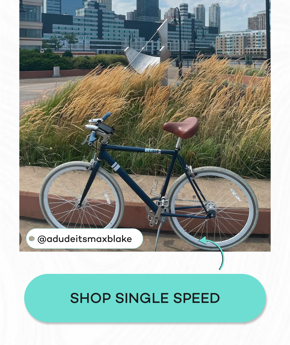 A blue single-speed bike parked near tall grass and modern buildings.