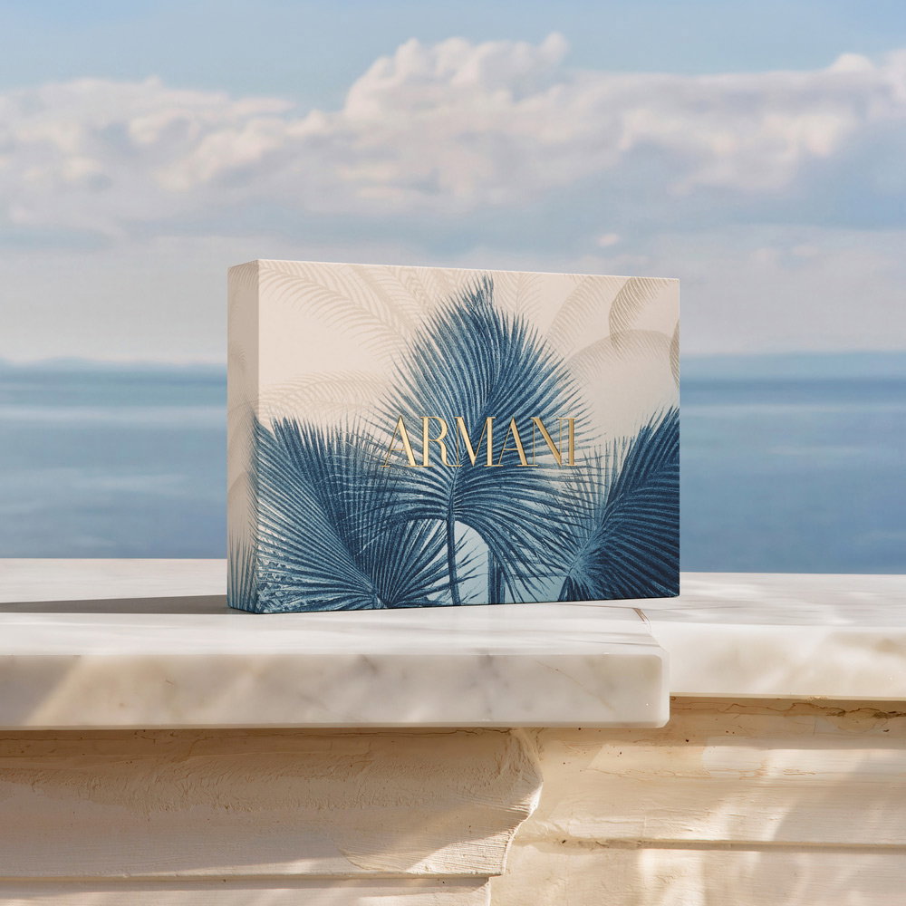 A luxury gift set presented on a marble ledge overlooking the sea. Includes a set of blue Armani playing cards, a blue-and-cream patterned hand towel, and a bottle of Acqua di Giò perfume.