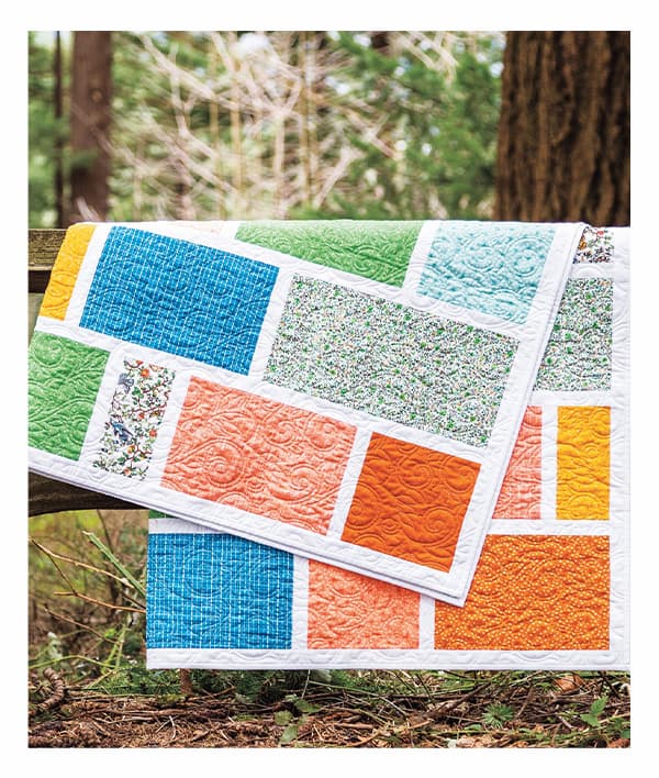 A colorful quilt with rectangular and square blocks is draped over a wooden fence. >>
