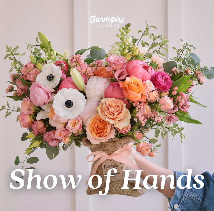 Farmgirl Flowers campaign image showing a large burlap-wrapped bouquet held by someone in a denim sleeve. The bouquet includes pink, peach, orange, white, and green flowers, with white text over the image that reads “Show of Hands.