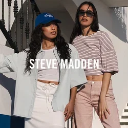 Zwei Frauen in legeren Sommeroutfits posieren neben einem weißen Gebäude mit blauer Verkleidung, einer Außentreppe und felsiger Küste unter klarem Himmel.
