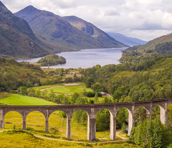 A scenic view of a stone viaduct spanning lush green hills and a tranquil lake surrounded by mountains.