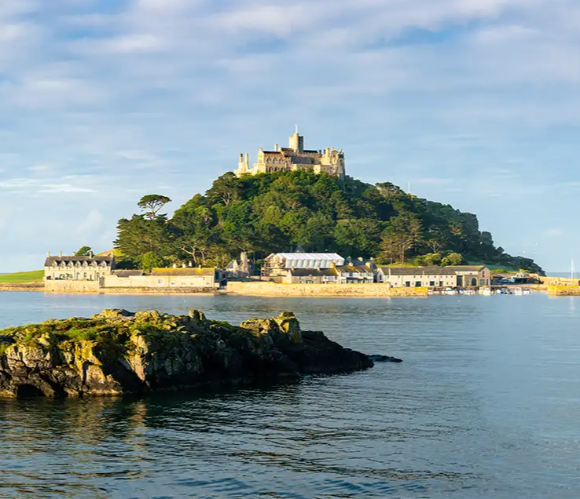A scenic view of a castle atop a green hill surrounded by water and rocky shoreline.
