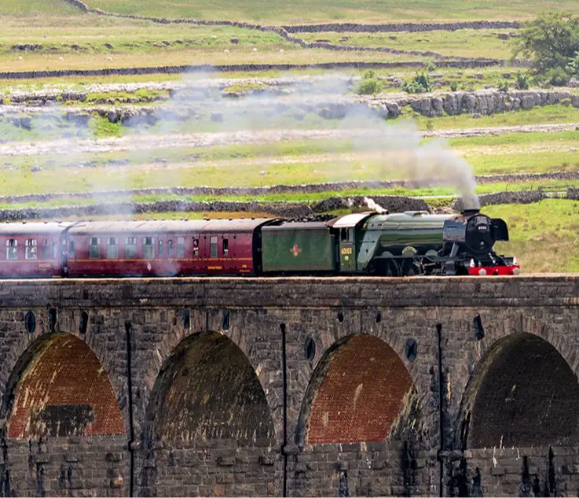 A steam locomotive crosses a stone viaduct, with green hills in the background.