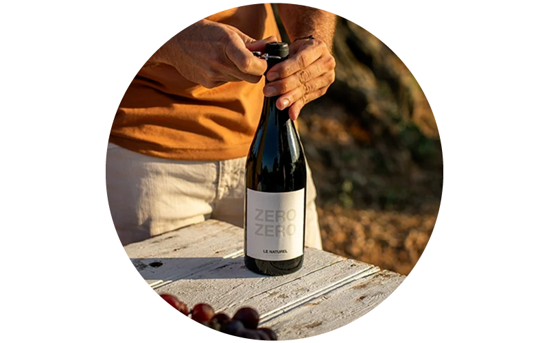 Hands opening a bottle of 'Zero Zero Le Naturel' natural wine on a white wooden table outdoors with grapes