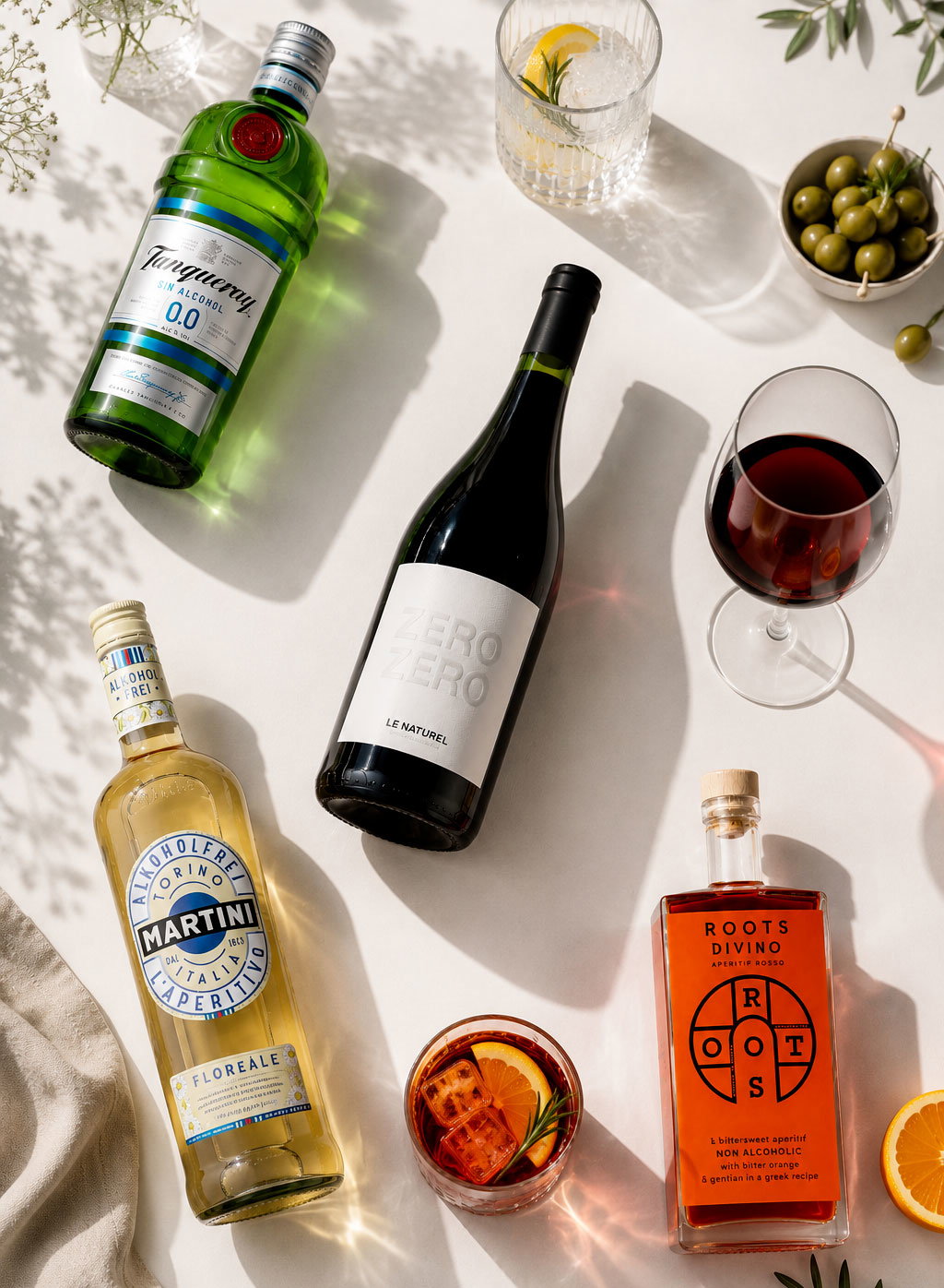 Flat lay of non-alcoholic spirits and aperitifs with a wine-style bottle, cocktail and red-wine glasses, orange slice and bowl of olives