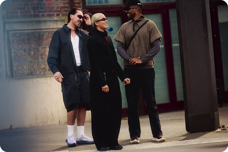 Three individuals standing outdoors smiling and laughing in casual, modern clothing, showcasing diverse fashion styles. The setting appears urban with a brick wall and glass doors in the background.