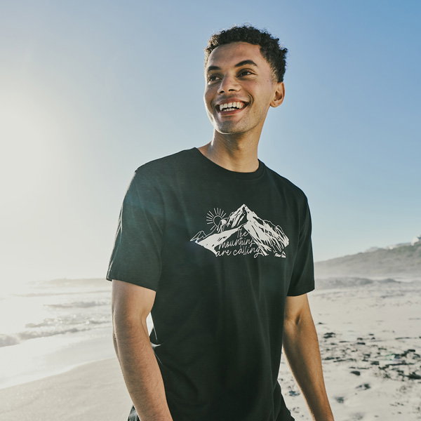Person standing on a sunny beach near the shoreline, wearing a black T-shirt with a white mountain graphic and text, looking off to the side with a smile; ocean waves and sandy coast visible in the background under clear blue sky.