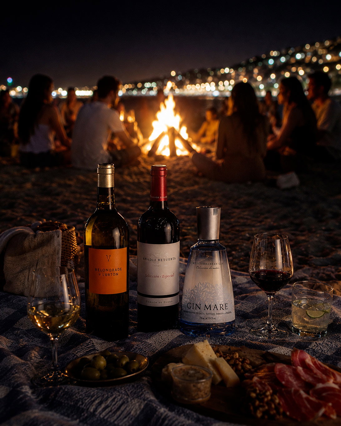 Night beach picnic with wine and gin bottles, glasses and charcuterie in foreground, friends around a bonfire and city lights