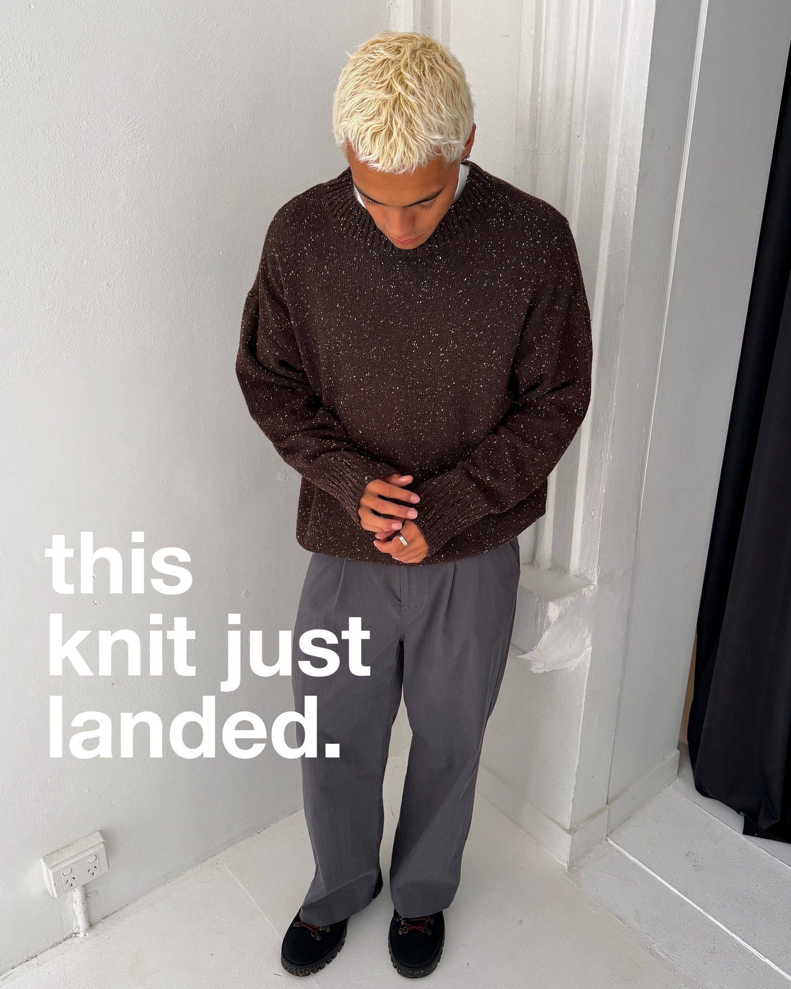 A medium shot shows a person with short, bleached blonde hair wearing a speckled brown knit sweater and grey trousers, standing with hands clasped in ...