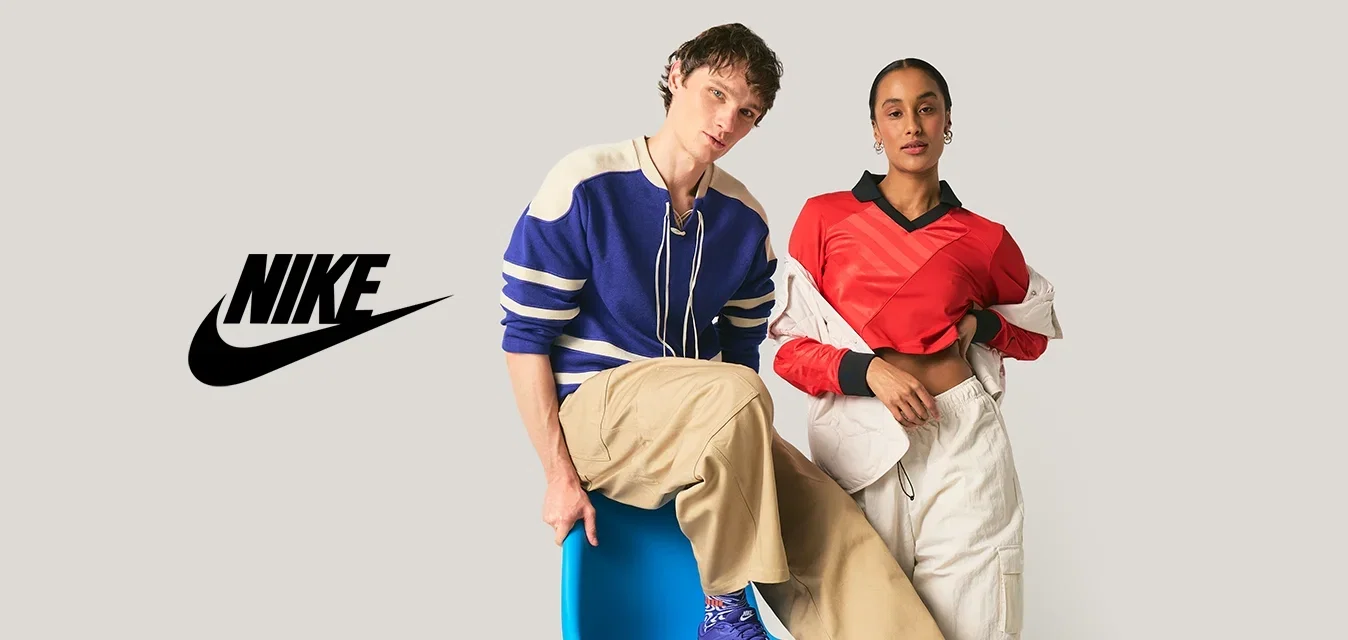 Zwei junge Erwachsene in lässigen Outfits posieren; einer sitzt auf einem blauen modernen Stuhl, der andere steht mit der Hand in der Tasche vor einem schlichten Hintergrund.