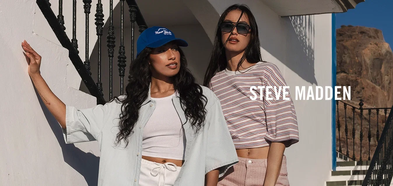 Zwei Frauen in legeren Sommeroutfits posieren neben einem weißen Gebäude mit blauer Verkleidung, einer Außentreppe und felsiger Küste unter klarem Himmel.