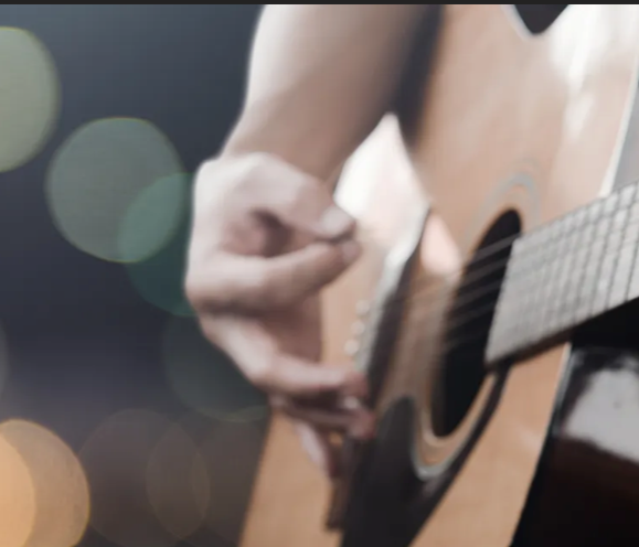 A close-up of a hand strumming an acoustic guitar with blurred lights in the background.