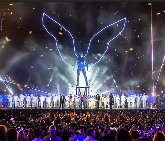 A large stage with a glowing angel figure and performers in white, surrounded by an enthusiastic crowd.
