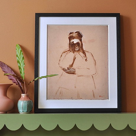 A black framed print of Edgar Degas' young woman holding a pair of field glasses design sits on a green shelf against a deep orange wall. - Prints and Postcards Collection.