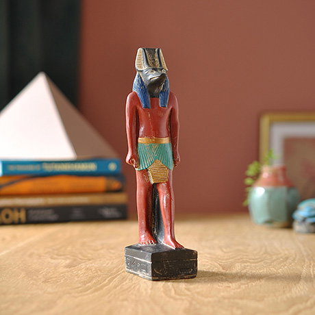 Hand-cast resin statue inspired by Anubis, the jackal-headed god of the underworld, displayed on a wooden surface with a pyramid-shaped silhouette in the distance. - Inspired by Ancient Egypt. 
