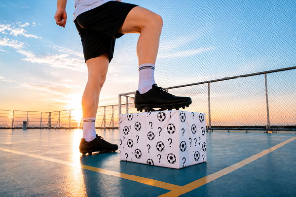 Bein in Sportschuh stellt einen Fuß auf einen würfelförmigen Kasten mit Fußballmuster auf einem Outdoor-Spielfeld bei Sonnenuntergang, Zaun im Hintergrund.