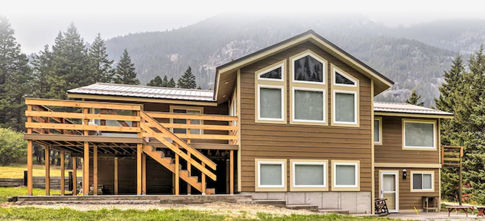 Wood-sided cabin with raised deck and stairs, set against mountains and evergreens.
