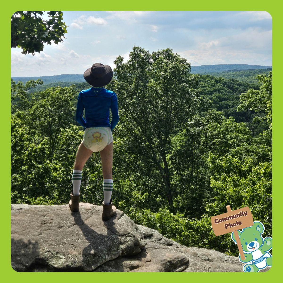 MischiefPup stands on a rocky hill looking over a forest. He is wearing an ABU Little Kings adult diaper. 