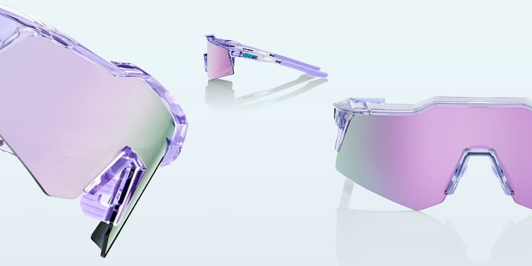 Close-up detail of the 100% Speedcraft XS "Translucent Lavender" sunglasses highlighting the bold blue 100% logo on the crystal frame, lavender rubber temple grip with honeycomb texture, and soft rose-tinted mirrored lens.