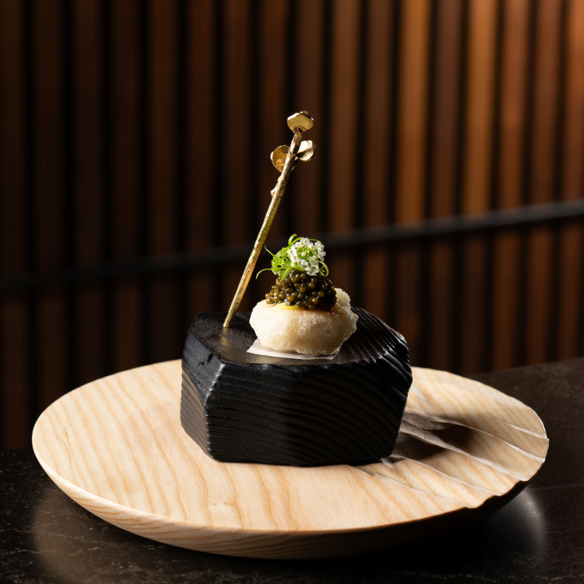 A refined gourmet appetizer is artfully presented on a light wooden plate. The food rests on a sculpted black block, creating a striking contrast in color and texture. A delicate vertical garnish rises from the center, topped with microgreens and a finely detailed topping. The background features warm, blurred vertical lines that emphasize the elegant restaurant setting.