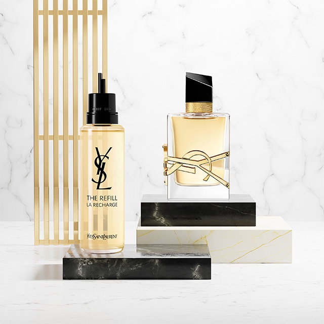 A YSL Libre fragrance bottle and its golden refill bottle displayed on marble and black stone pedestals.