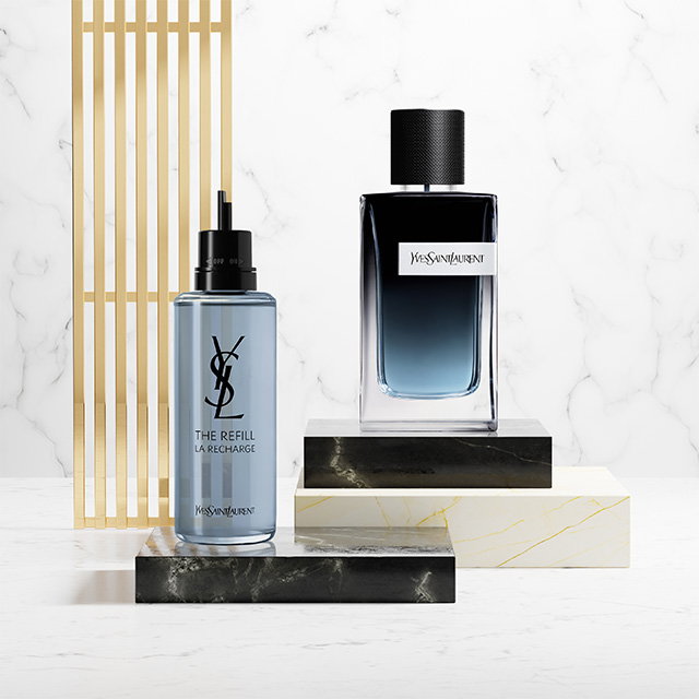 A blue YSL Y fragrance bottle and its light blue refill bottle displayed on marble and black stone pedestals.