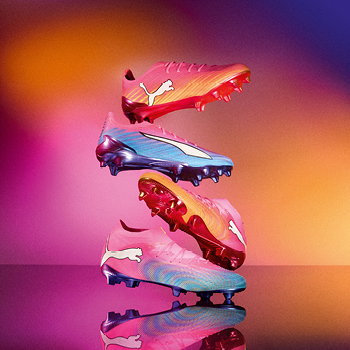 Vibrant stacked Puma football boots in pink, blue and gold on a reflective surface with a warm gradient background