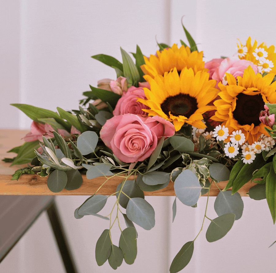 Close-up image of the Summer Fridays bouquet featuring yellow sunflowers, pink roses, small white daisies, pink alstroemeria, greenery, and eucalyptus arranged against a light background.