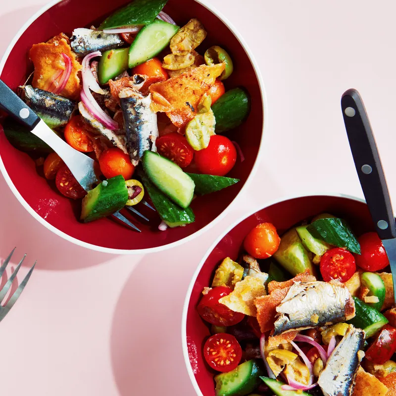 Broken up oil-packed sardines in three bowls with chopped preserved lemons, Persian cucumbers, cherry tomatoes, red onions, mixed olives, and salted pita chips.