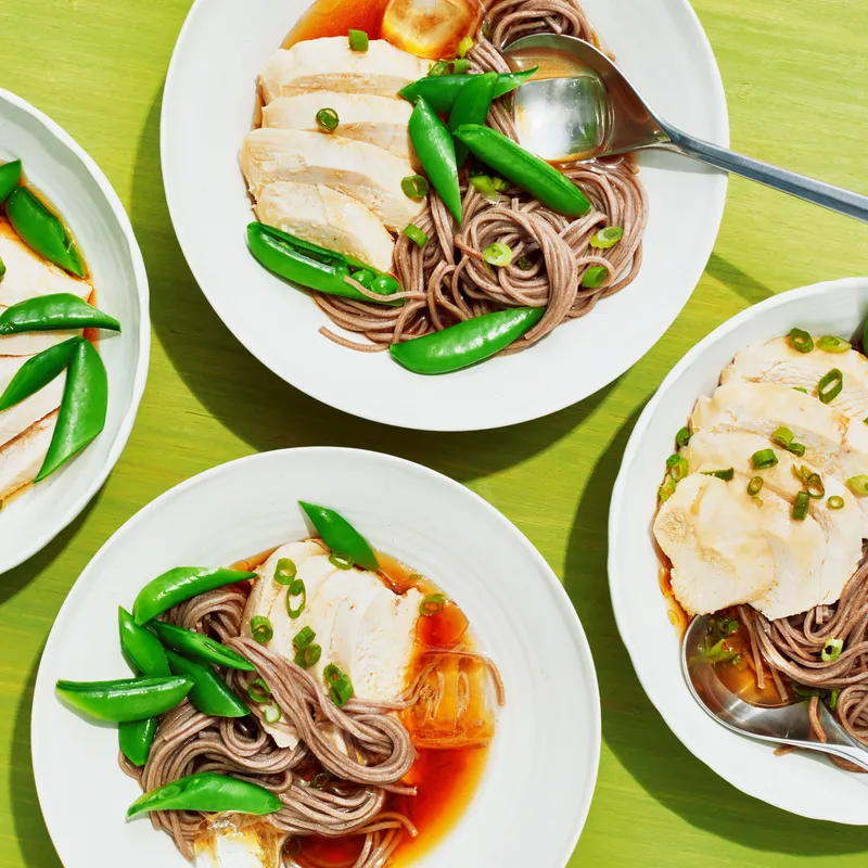 Four bowls of boiled and sliced chicken breast, soba, snap peas, miso paste, lemon juice, and soy sauce topped with chopped scallions, sesame oil, and ice cubes.
