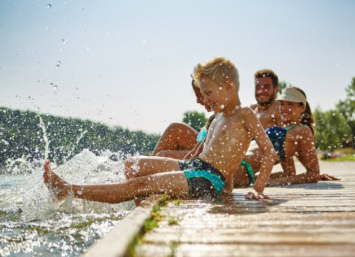 Familie aus Mann, Frau, Tochter und Sohn sitzen an einem Steg und Planschen mit den Beinen im Wasser. Jetzt sparen.