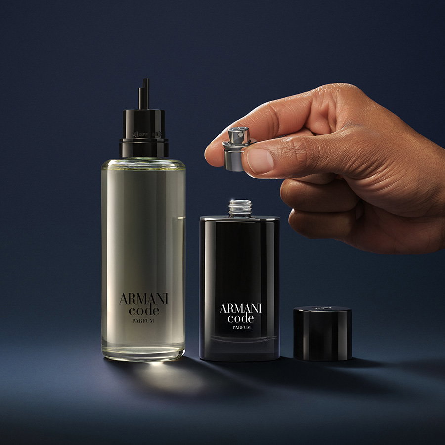 A hand placing a sleek black Armani Code bottle next to its large glass refill container against a dark blue background.