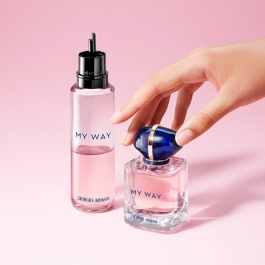A hand placing a pink My Way perfume bottle with its iconic blue cap next to a tall refill bottle on a soft pink background.