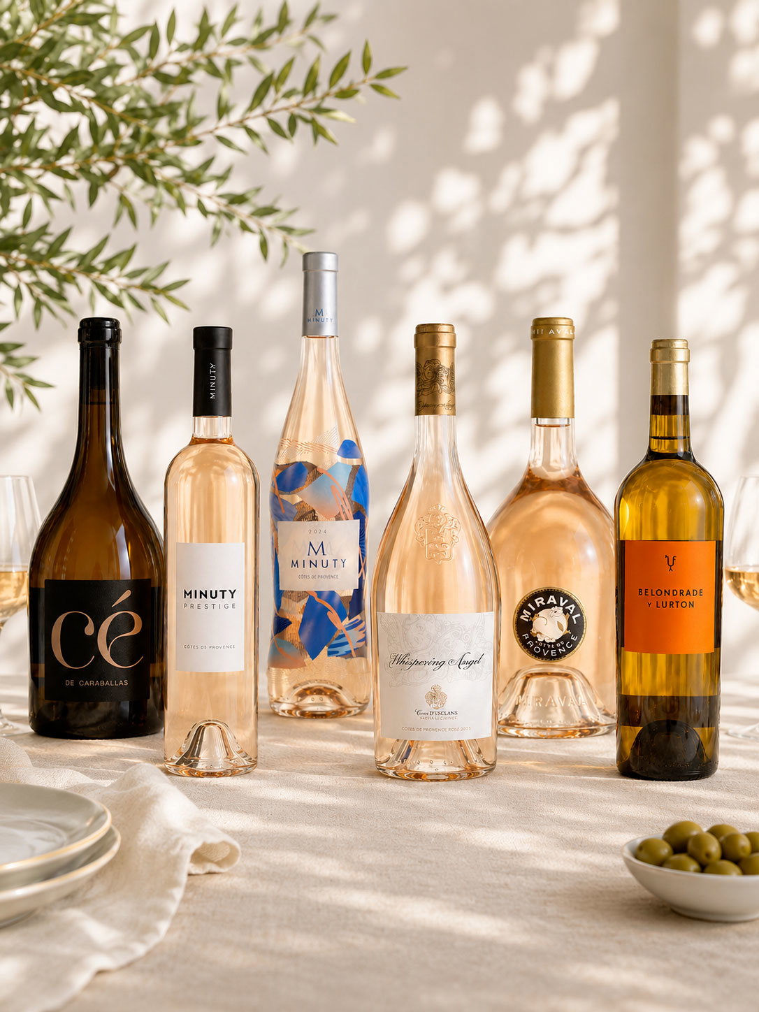 Six assorted rosé wine bottles and a white wine on a sunlit table with olives, plates and dappled shadows
